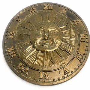 May include: A circular bronze-toned clock face featuring a central embossed sun with a human face and radiating rays. The outer edge displays raised Roman numerals from I to XII, with the names of the twelve months inscribed between the numerals.