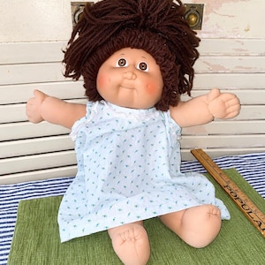 May include: A doll with brown yarn hair, rosy cheeks, and brown eyes, wearing a light blue sleeveless dress with a small blue and green floral pattern. A wooden ruler is visible next to the doll, showing measurements in inches.
