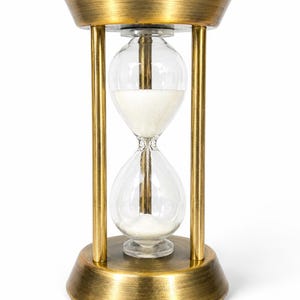 May include: An hourglass featuring a brushed gold metal frame with a circular base and top. The central glass bulb contains fine white sand, with a portion of the sand currently resting in the bottom chamber, marking the passage of time.