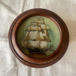 May include: A small, round wooden frame encases a painting of a tall ship with billowing sails. The ship is depicted in shades of brown, black, and white against a light green background. A smaller ship is visible in the distance.