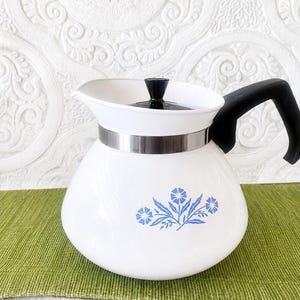 May include: A white teapot with a rounded body featuring a blue floral pattern. The teapot has a black handle, a black lid knob, and a silver-toned metal band encircling the upper section just below the wide, open spout and rim.