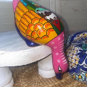 May include: A hand-painted ceramic duck figurine. The duck features a pink head and tail, a yellow body with orange detailing, and a blue base. The body is decorated with a landscape scene. The figurine is on a white surface.