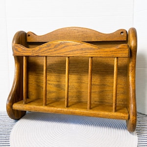 May include: A wall-mounted wooden magazine rack featuring a medium brown oak finish. The rack has a curved top edge, solid side panels, and a front section composed of four vertical spindles that create an open storage space for holding items.