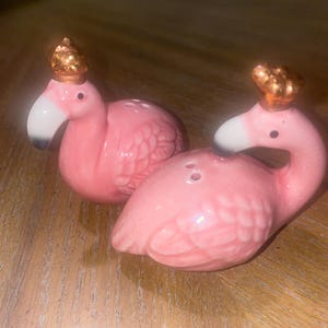 May include: A pair of pink ceramic flamingo salt and pepper shakers. Each flamingo has a white beak, black eye, and a gold-colored crown. The shakers are designed with a textured feather pattern.