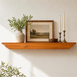 May include: A wooden shelf with a warm, orange-brown finish is mounted on a white wall. It holds a framed landscape painting, a beige vase with green foliage, a small wooden box, and two brass candlesticks with white candles.