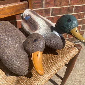 Vintage Flambeau mallard duck decoy pair displayed outdoors on a woven rush seat chair against a red brick wall, featuring a realistic drake with deep green head, white neck ring, gray body and blue wing accent alongside a brown hen with warm feather tones, both molded plastic hunting decoys with detailed feather texture and aged patina, functional floating waterfowl decoys with weighted keel bases, styled rustic cabin lodge decor, lake house interior, Ralph Lauren inspired sporting aesthetic