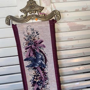 Top section of signed Glenda Turley textile wall hanging showing ornate brass style metal mount hardware curved decorative form and woven floral bouquet ribbon and bird motif in plum burgundy blue and sage tones framed by twisted cord trim showcasing fiber art texture dimensional threads and Victorian inspired romantic traditional home decor style