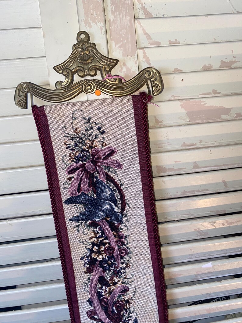 Top section of signed Glenda Turley textile wall hanging showing ornate brass style metal mount hardware curved decorative form and woven floral bouquet ribbon and bird motif in plum burgundy blue and sage tones framed by twisted cord trim showcasing fiber art texture dimensional threads and Victorian inspired romantic traditional home decor style