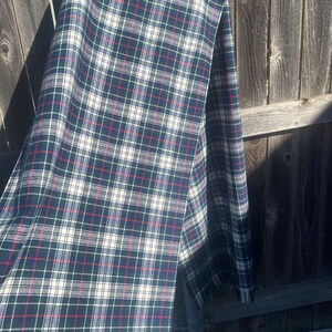 May include: A folded plaid scarf in shades of dark green, navy, red, and white. The scarf is draped over a wooden fence, showcasing its intricate pattern. The fabric appears to be soft and suitable for cooler weather.