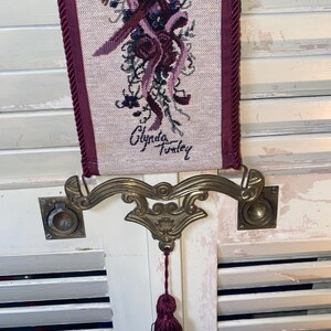 Signed Glenda Turley woven textile wall hanging featuring floral bouquet ribbon and bird motif in rich jewel tones including burgundy plum blue and sage framed by corded trim with ornate brass tone metal mount and decorative tassel pull showing vertical fiber art format vintage Victorian inspired home decor piece with detailed woven threads and dimensional texture suitable for traditional cottage or romantic interiors