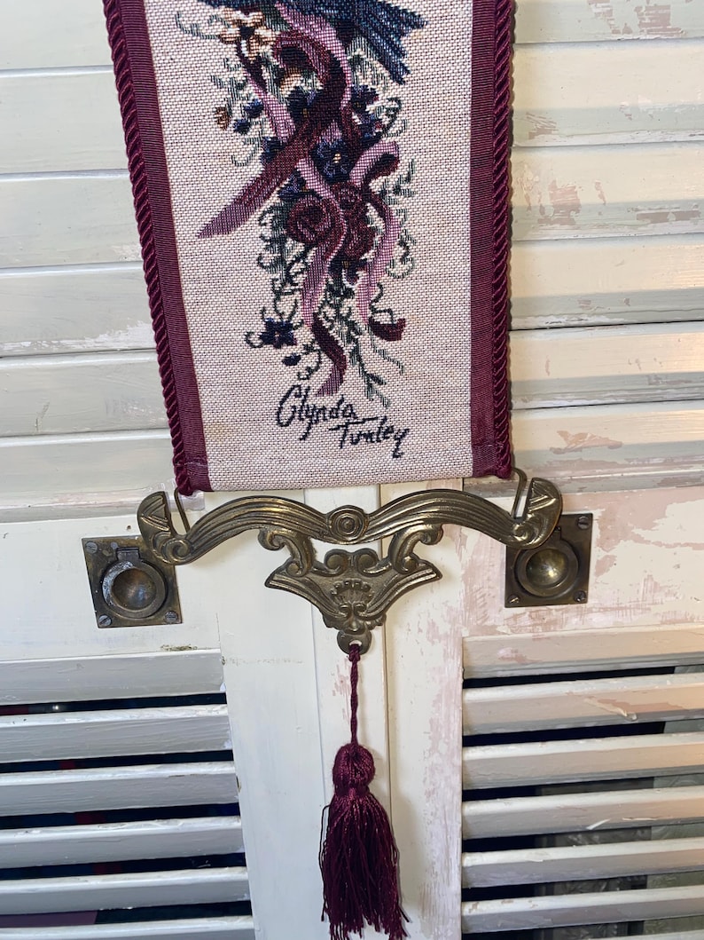 Signed Glenda Turley woven textile wall hanging featuring floral bouquet ribbon and bird motif in rich jewel tones including burgundy plum blue and sage framed by corded trim with ornate brass tone metal mount and decorative tassel pull showing vertical fiber art format vintage Victorian inspired home decor piece with detailed woven threads and dimensional texture suitable for traditional cottage or romantic interiors