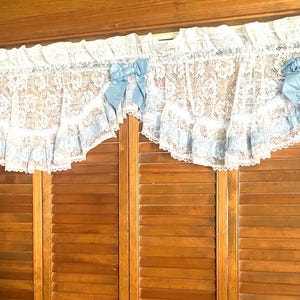 May include: A white lace window valance featuring a floral pattern, ruffled edges, and three light blue satin bows attached across the top. The valance is gathered along the rod pocket and hangs with a scalloped hemline over a wooden door.