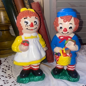 May include: Two ceramic figurines of Raggedy Ann and Andy. Raggedy Ann wears a yellow dress, white apron, and holds a red apple. Andy wears a blue outfit, a red bow tie, and carries a picnic basket with a sandwich. Both have red yarn hair.