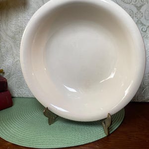 May include: A cream-colored, round ceramic bowl with a wide rim. The bowl is set on a brass stand and a green placemat. The background features a patterned wallpaper and a wooden surface.