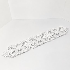 May include: A long, narrow, white decorative molding piece featuring an intricate, symmetrical relief pattern of scrolling acanthus leaves and floral motifs. The object has a matte finish and a repeating design that spans its entire length.