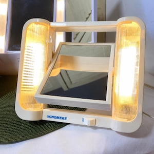 May include: A cream-colored Windmere lighted vanity mirror with a rectangular tilting center mirror and two vertical ribbed light panels on either side. The base features a small white power switch labeled with ON and OFF settings.