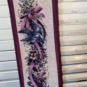 Close view of woven floral ribbon and bird design in Glenda Turley tapestry showing detailed thread work layered texture jewel tone color palette burgundy plum blue and green framed by corded trim emphasizing fiber art craftsmanship vintage textile wall decor and romantic Victorian inspired interior styling element