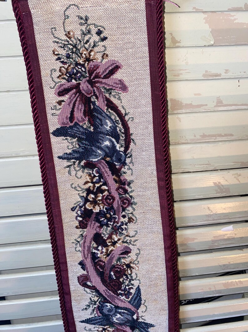 Close view of woven floral ribbon and bird design in Glenda Turley tapestry showing detailed thread work layered texture jewel tone color palette burgundy plum blue and green framed by corded trim emphasizing fiber art craftsmanship vintage textile wall decor and romantic Victorian inspired interior styling element