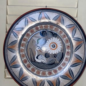 May include: A decorative ceramic plate with a hand-painted design. The plate features a central image of a white flower and a bird, surrounded by a border of stylized leaves and floral patterns in shades of blue, orange, and white. The rim is a dark red.