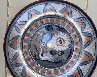 Puede incluir: Un plato de cerámica decorativo con un diseño pintado a mano. El plato presenta una imagen central de una flor blanca y un pájaro, rodeada por un borde de patrones florales y geométricos en tonos de azul, naranja y blanco.