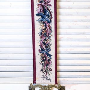 Vertical Glenda Turley signed tapestry wall hanging with Victorian floral ribbon and bird design in plum burgundy blue and sage tones woven into textured textile fabric framed by twisted cord trim mounted in ornate brass style decorative hardware at top and bottom with deep burgundy tassel pull elegant vintage fiber art wall decor piece displayed against neutral backdrop showing full length proportions intricate woven detail and classic traditional romantic style