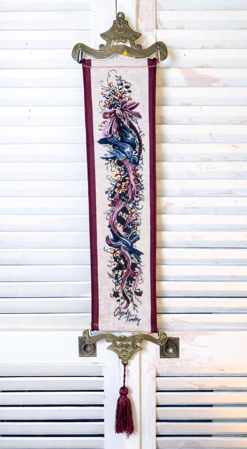 Vertical Glenda Turley signed tapestry wall hanging with Victorian floral ribbon and bird design in plum burgundy blue and sage tones woven into textured textile fabric framed by twisted cord trim mounted in ornate brass style decorative hardware at top and bottom with deep burgundy tassel pull elegant vintage fiber art wall decor piece displayed against neutral backdrop showing full length proportions intricate woven detail and classic traditional romantic style