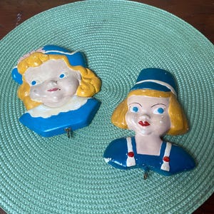 May include: Two vintage ceramic wall hooks shaped like children's faces. One has blonde hair and a blue hat and collar, the other has blonde curls and a blue hat and collar. Both have a hook for hanging items.