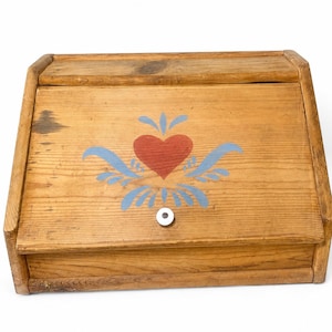 May include: A rectangular wooden bread box with a slanted front lid featuring a painted red heart and blue decorative flourishes. The natural wood grain is visible across the surface, and a small, round, white knob is centered on the front panel.