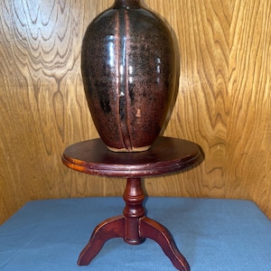 May include: A dark brown ceramic vase with a glossy finish and a unique, textured design. The vase is sitting on a small, wooden pedestal.