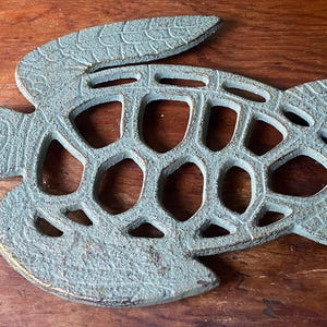 May include: A decorative, cast-iron turtle-shaped trivet with a distressed, turquoise finish. The trivet features a detailed design with open spaces, and is placed on a wooden surface. The turtle's shell has a textured pattern.