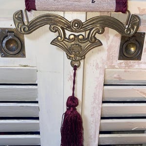 Ornate brass tone bottom hardware of Glenda Turley tapestry wall hanging featuring curved scrollwork design metal mount and attached deep burgundy tassel pull with woven textile panel above showing floral ribbon and bird pattern in jewel tones vintage decorative wall art with traditional Victorian aesthetic