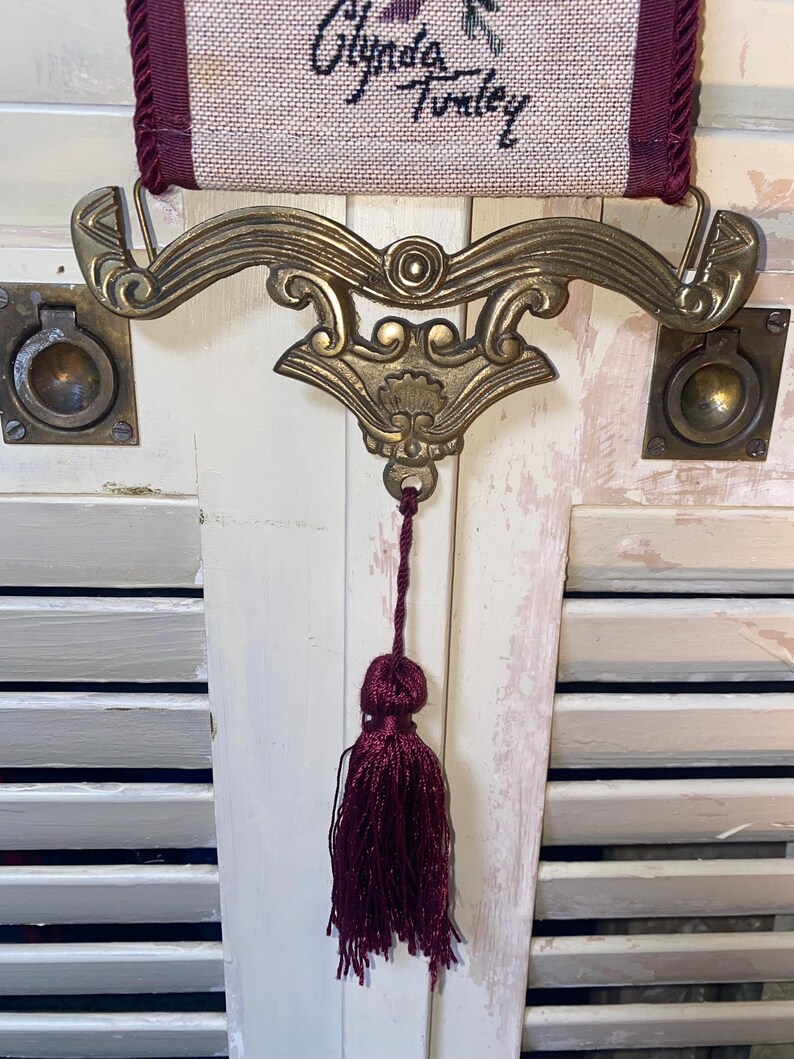 Ornate brass tone bottom hardware of Glenda Turley tapestry wall hanging featuring curved scrollwork design metal mount and attached deep burgundy tassel pull with woven textile panel above showing floral ribbon and bird pattern in jewel tones vintage decorative wall art with traditional Victorian aesthetic