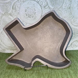 May include: A metallic, light brown tray shaped like the state of Texas. The tray features a smooth, matte finish with raised edges and a hollow center. It is supported by a thin, silver-colored wire stand that holds the tray in an upright position.