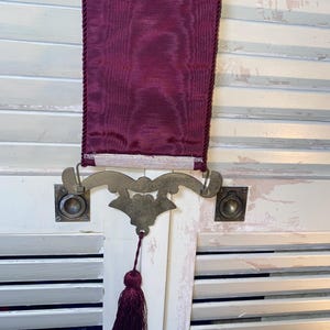 Lower portion of Glenda Turley tapestry wall art showing woven bird and floral ribbon details in textured textile fabric with burgundy cord trim ornate brass finish bottom mount hardware and deep burgundy tassel pull highlighting decorative metal scrollwork design fiber art craftsmanship and vintage traditional wall decor aesthetic with rich jewel tone color palette and intricate woven thread pattern