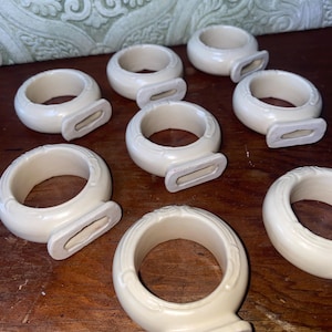 May include: A collection of off-white ceramic napkin rings. Each ring has a rounded shape with a decorative base and a rectangular opening. The rings are arranged on a wooden surface.