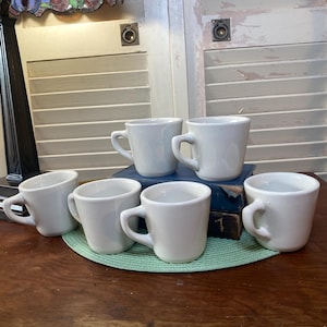 May include: A collection of six white ceramic coffee cups with curved handles. The cups are arranged on a wooden surface, with some resting on a green mat and others on a stack of books. The background includes a lamp and a white shuttered door.