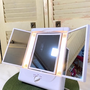 May include: A white tri-fold vanity mirror with integrated vertical LED lighting strips. The base features a central rotary dial with settings labeled OFF, EVENING, HOME, OFFICE, and DAY. The mirror panels are adjustable for multiple viewing angles.