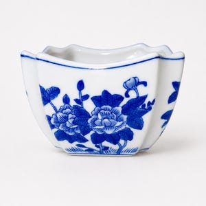 May include: A small white ceramic bowl with a scalloped rim and a blue floral pattern featuring detailed blossoms and leaves painted on the exterior. The bowl has a curved, multi-sided shape and a smooth, glossy finish throughout the surface.