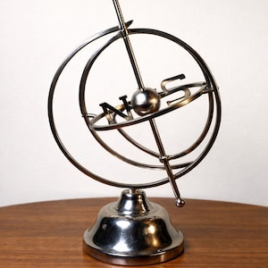 May include: A polished metal decorative globe with a compass design. The globe features a spherical center with a vertical rod and intersecting rings, mounted on a flared base. The letters N and S are visible.