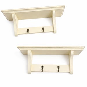 May include: Two off-white wall shelves with a flat top surface and a lower horizontal bar. Each shelf features two dark metal hooks attached to the lower bar, supported by two side brackets that connect the top shelf to the bottom bar structure.