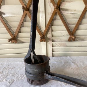 May include: An antique, dark metal potato ricer with a perforated base and a long handle. The ricer has a cylindrical shape with a handle on the side and a long lever for pressing. The kitchen tool is likely vintage and shows signs of wear.