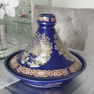 Puede incluir: Una tagine de cerámica azul con un diseño de pavo real dorado y blanco. La tagine tiene una base redonda y una tapa cónica.