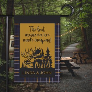 Op de afbeelding: Een zwarte en gouden tuin vlag met een eland silhouet en de tekst "The best memories are made camping!" en "Linda & John".