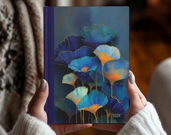 May include: A hardcover journal with a floral design. The cover features blue and orange flowers with gold accents against a dark blue background. The journal has a dark blue spine and is held by a person wearing a white sweater.
