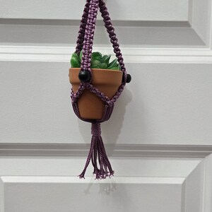 Peut inclure: Une petite plante en pot suspendue à un porte-plante en macramé violet avec des perles noires. Le porte-plante est accroché à un crochet blanc sur un mur blanc.
