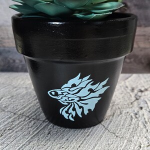 May include: A black terracotta pot with a blue fish design, filled with a green succulent plant.