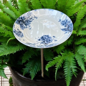 May include: A blue and white porcelain bird bath with a floral design. The bird bath is on a metal stake and is surrounded by green ferns.