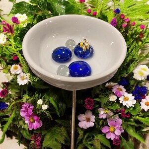 May include: A white ceramic bird bath filled with blue glass stones and clear glass marbles. A small gold-colored bee figurine sits atop one of the blue stones. The bird bath is surrounded by a vibrant arrangement of colorful flowers and greenery.