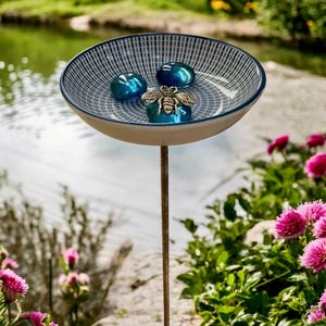 May include: A decorative bird bath with a ceramic bowl featuring a blue and white pattern. The bowl holds three blue glass stones and a silver bee figurine. The bird bath is mounted on a metal rod, set against a backdrop of a pond and pink flowers.