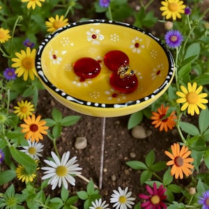 May include: A yellow ceramic bird bath with a black and white rim, decorated with white flowers. The bird bath holds three red glass beads, one with a gold bee. The bird bath is on a metal stake, surrounded by colorful flowers.